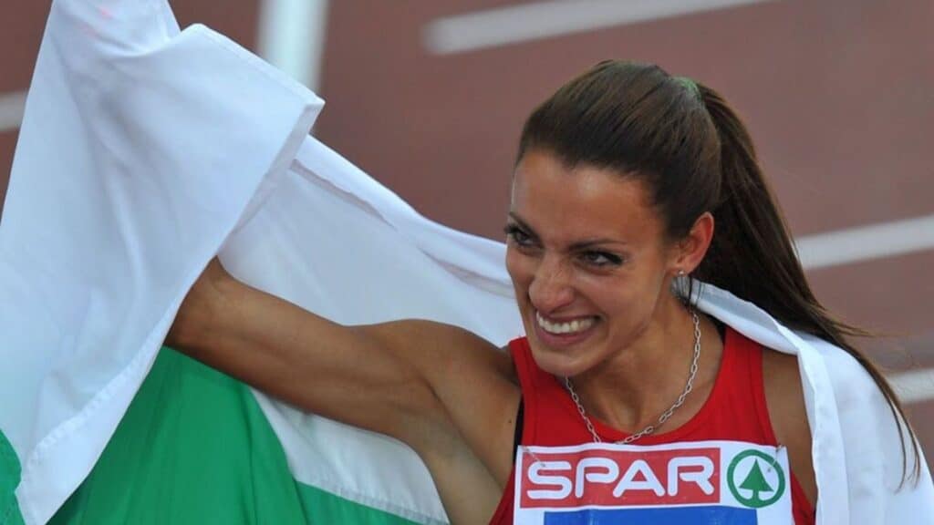 Ivet Lalova-Collio holding up the Bulgarian flag and smiling, wearing a red competition outfit during a track event.