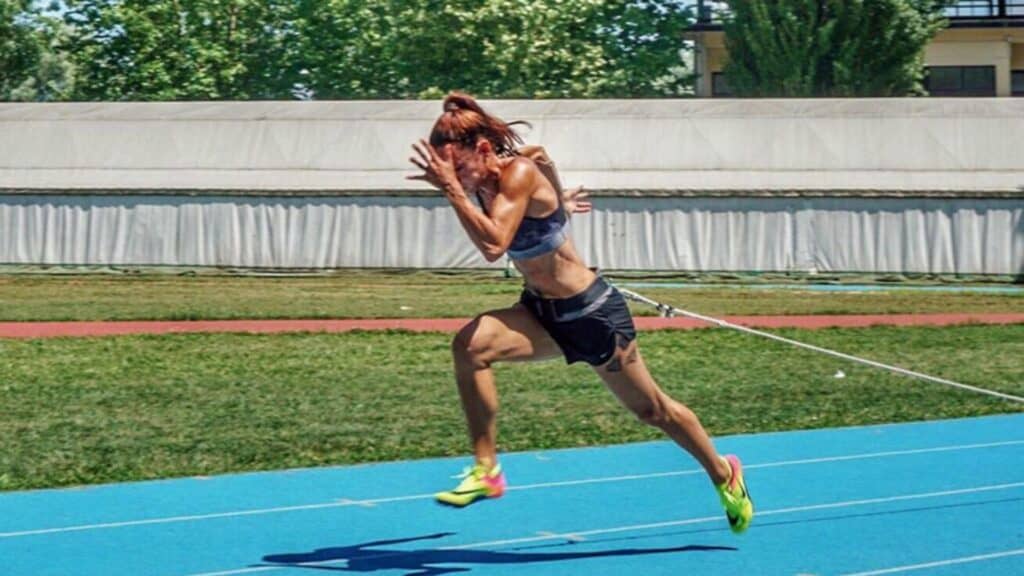 Ivet Lalova-Collio performing sprint training with a resistance band on a blue track, pushing forward with intensity.