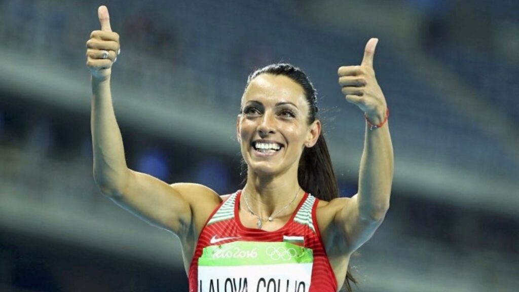 Ivet Lalova-Collio smiling and giving a double thumbs-up gesture during a competition, wearing a red competition outfit.