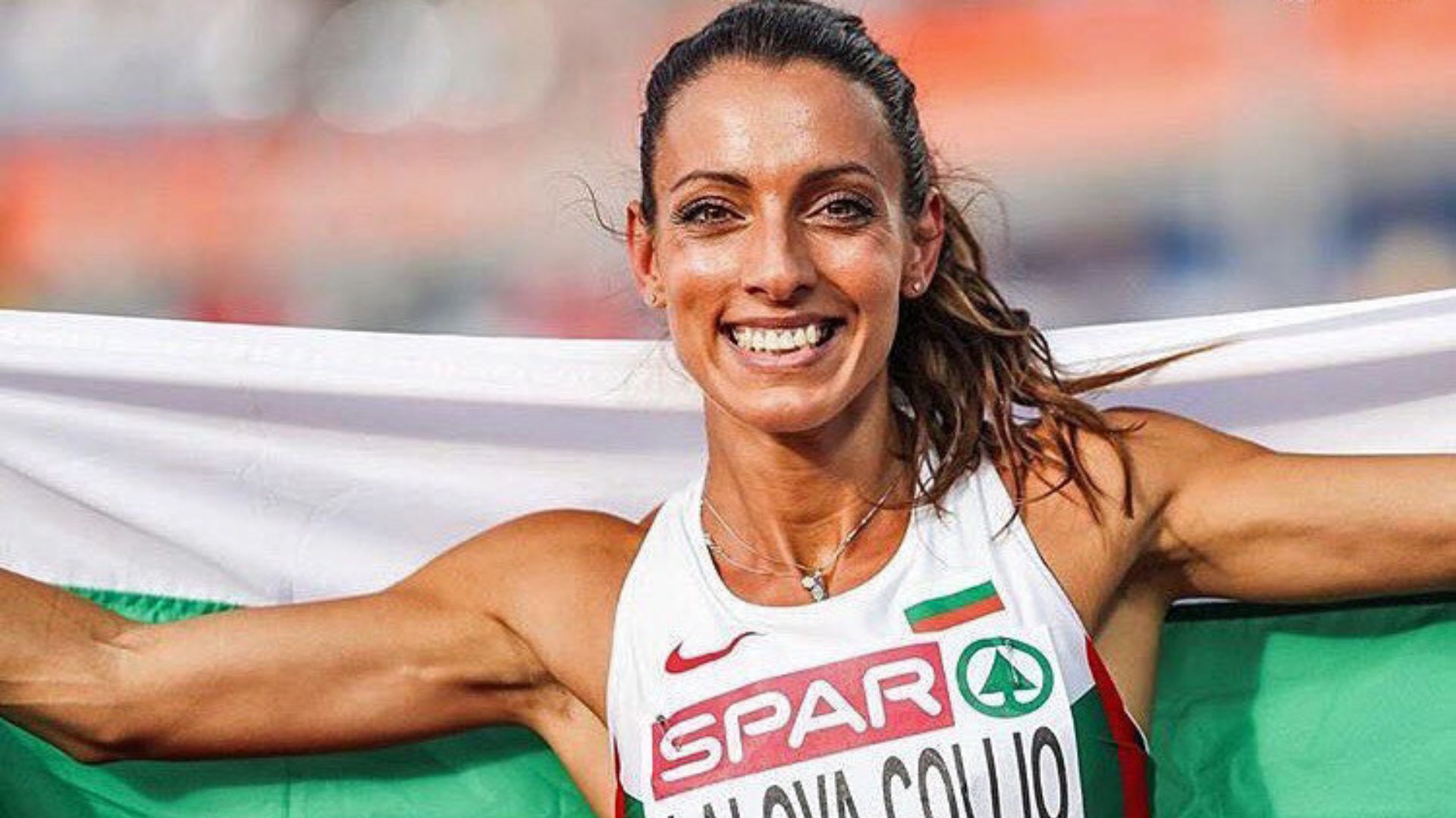 Ivet Lalova-Collio smiling broadly and holding the Bulgarian flag behind her after a successful race, wearing a white racing top.