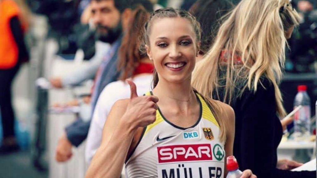 Laura Müller smiling and giving a thumbs-up while holding a water bottle, wearing a competition outfit with the German national insignia.