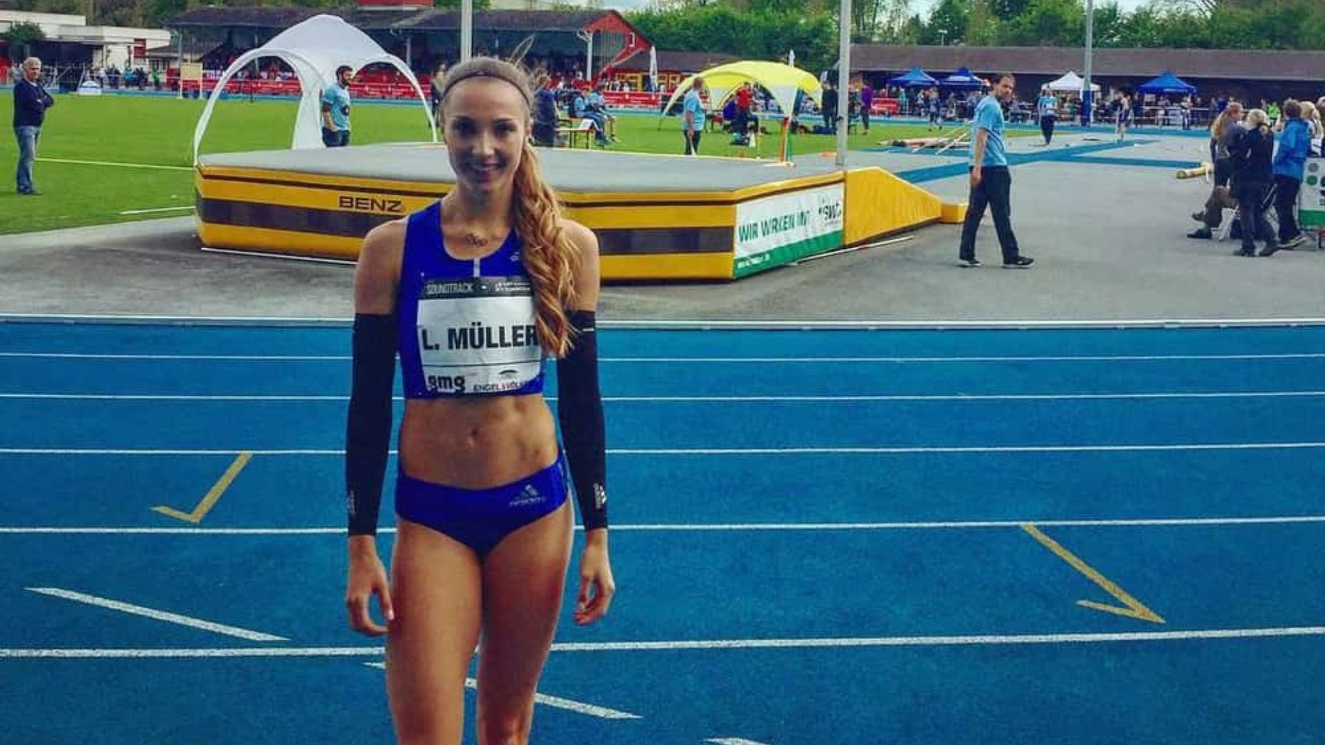 Athlete Laura Müller standing on a blue track in athletic gear with a competition bib, with a high jump setup and spectators in the background.