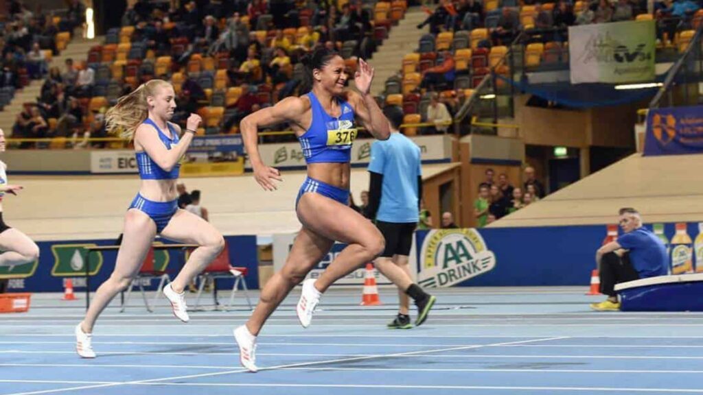 Athlete Naomi Sedney sprinting during an indoor track competition, leading ahead of competitors in blue athletic gear.