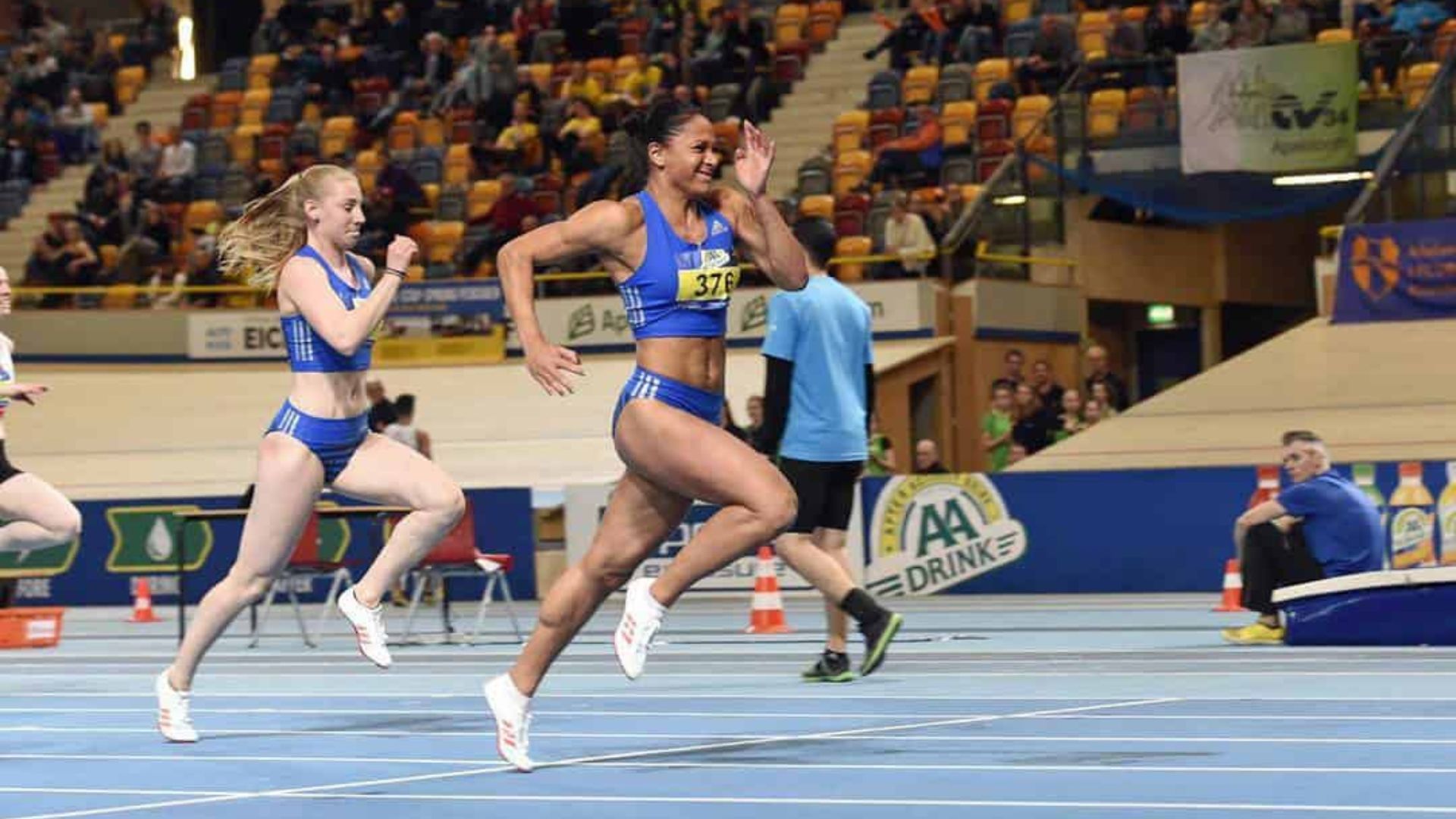 Athlete Naomi Sedney sprinting during an indoor track competition, leading ahead of competitors in blue athletic gear.