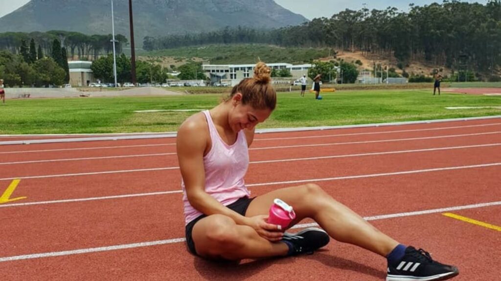 Naomi Sedney sitting on a red track holding a pink bottle, taking a break with a mountainous landscape in the background.