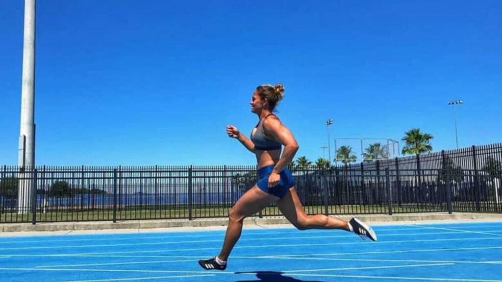 Naomi Sedney running on a blue outdoor track under a clear blue sky, wearing athletic shorts and a sports bra.