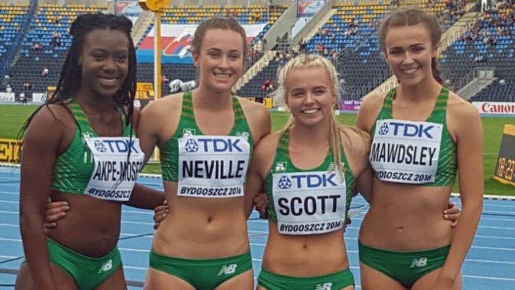 Sharlene Mawdsley posing with her relay teammates in green uniforms on a track, smiling after a competition with stadium seating in the background.