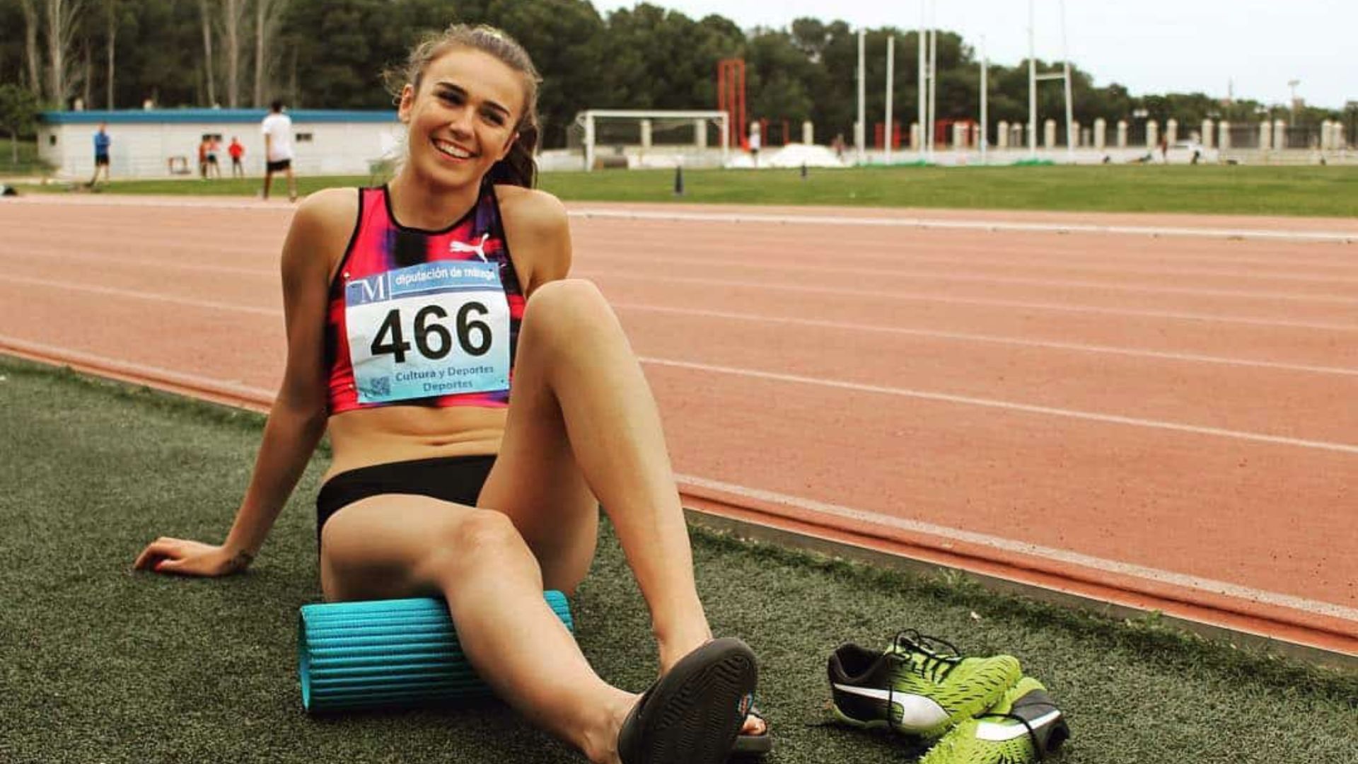 Sprint athlete Sharlene Mawdsley smiling and sitting on the grass by a track, with a foam roller and green spikes beside her after a race.